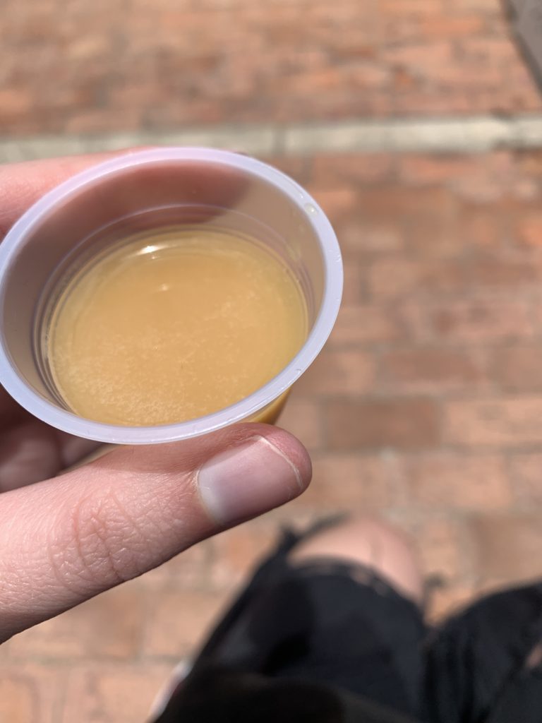 A thumb and pointer finger holding a small plastic cup filled with chicha, a brown and yellow fermented corn drink